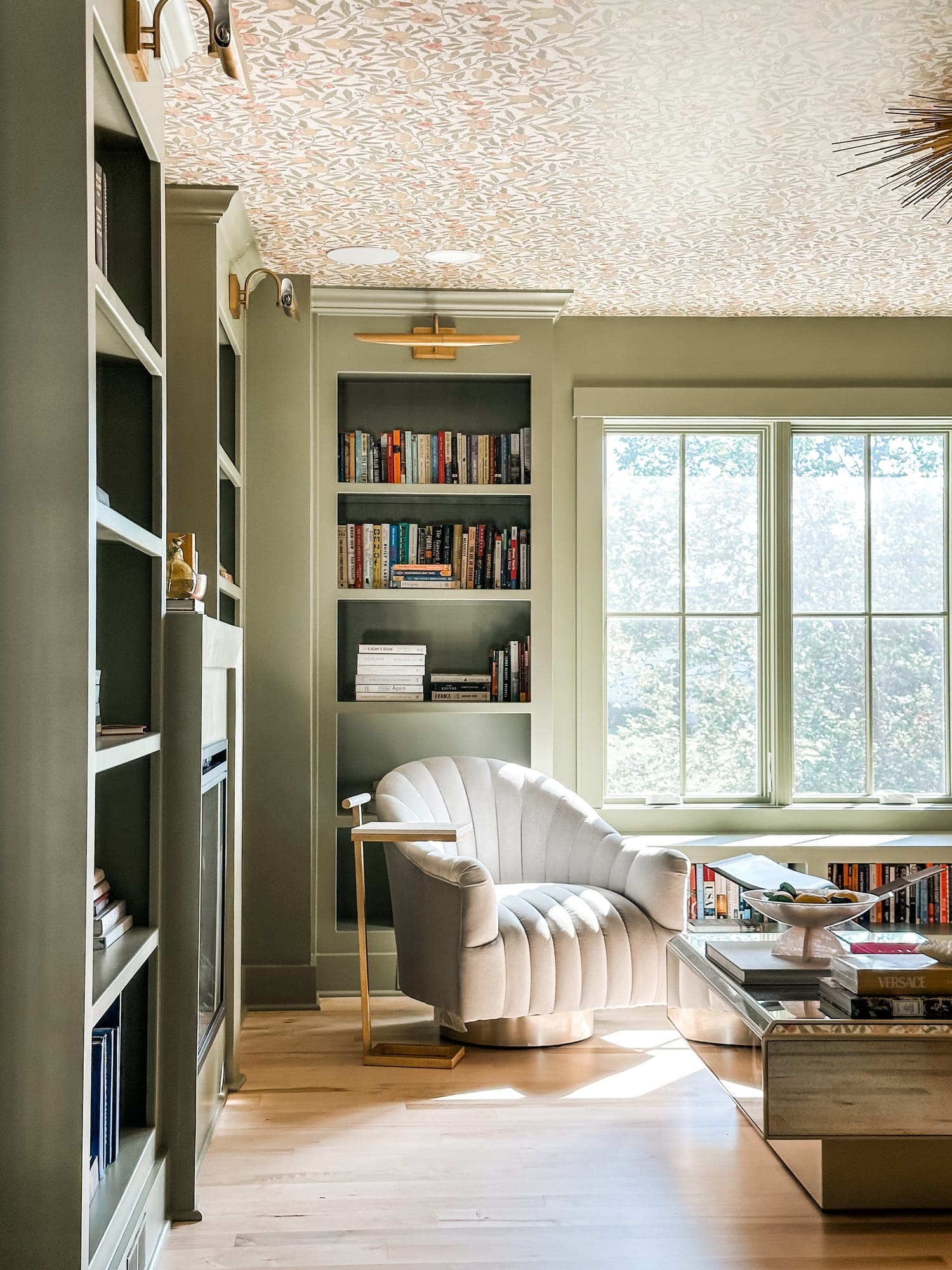 Living room with sage-green built-in bookshelves, floral wallpapered ceiling, and a cream velvet swivel chair, designed by LaMira Interiors in Overland Park, Kansas